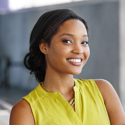 A woman with a smile, wearing a yellow top and standing in front of a building.
