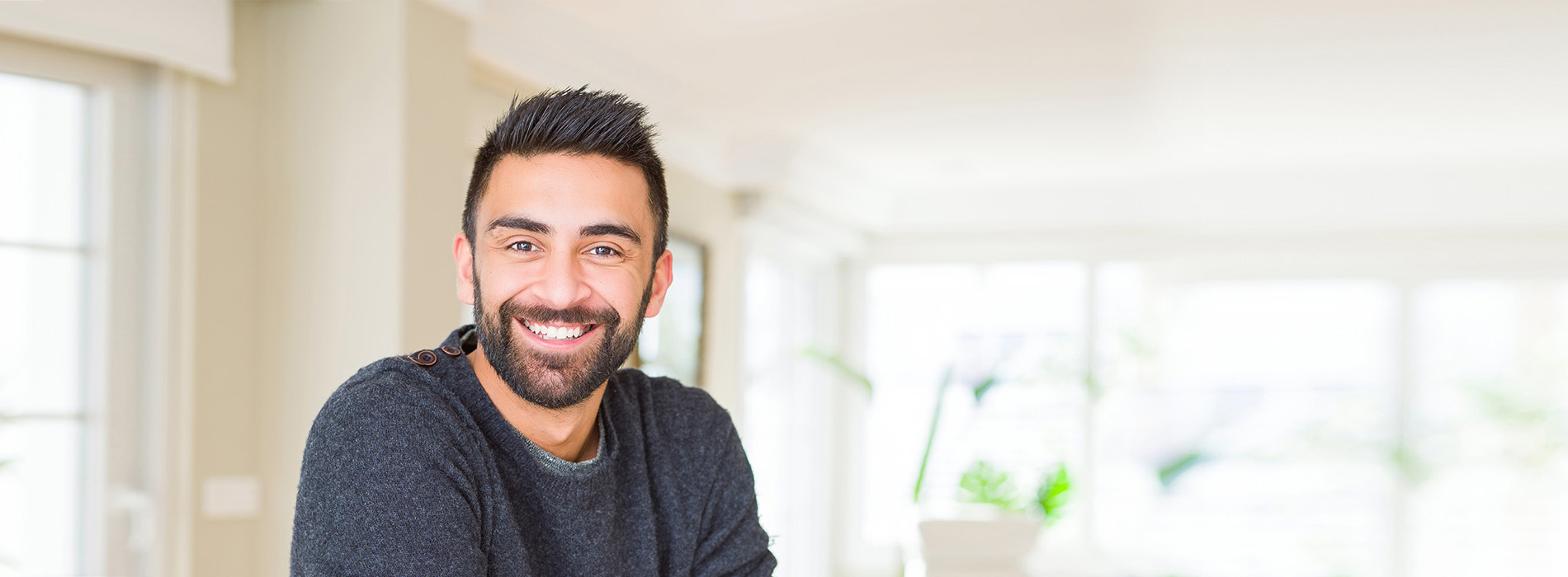 The image is a photograph of a man with facial hair, smiling at the camera. He appears to be indoors, possibly in a home or office setting, with a blurred background that suggests a domestic environment.